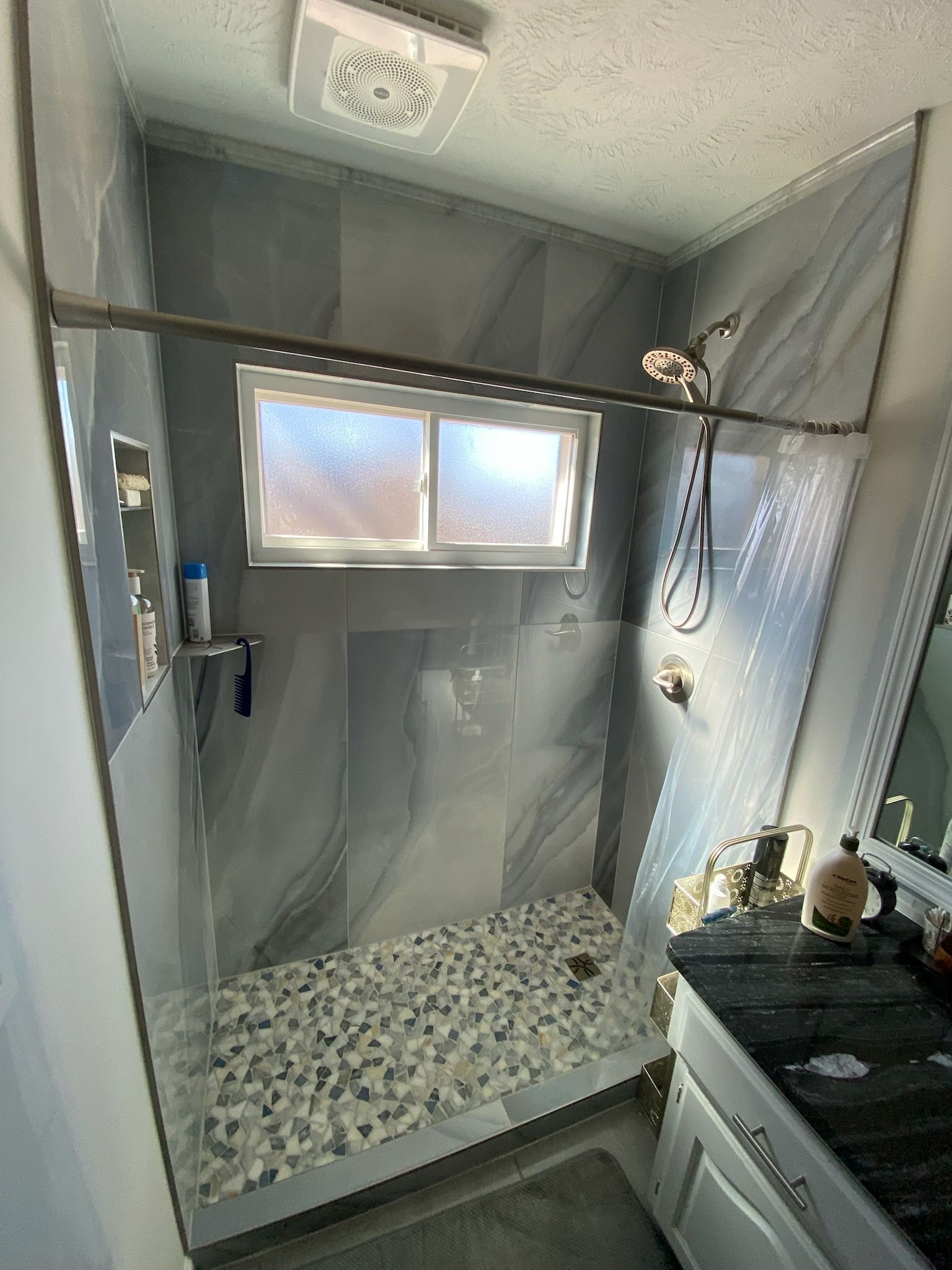 Shower with gray and white tile, pebble floor, glass door, and silver fixtures.