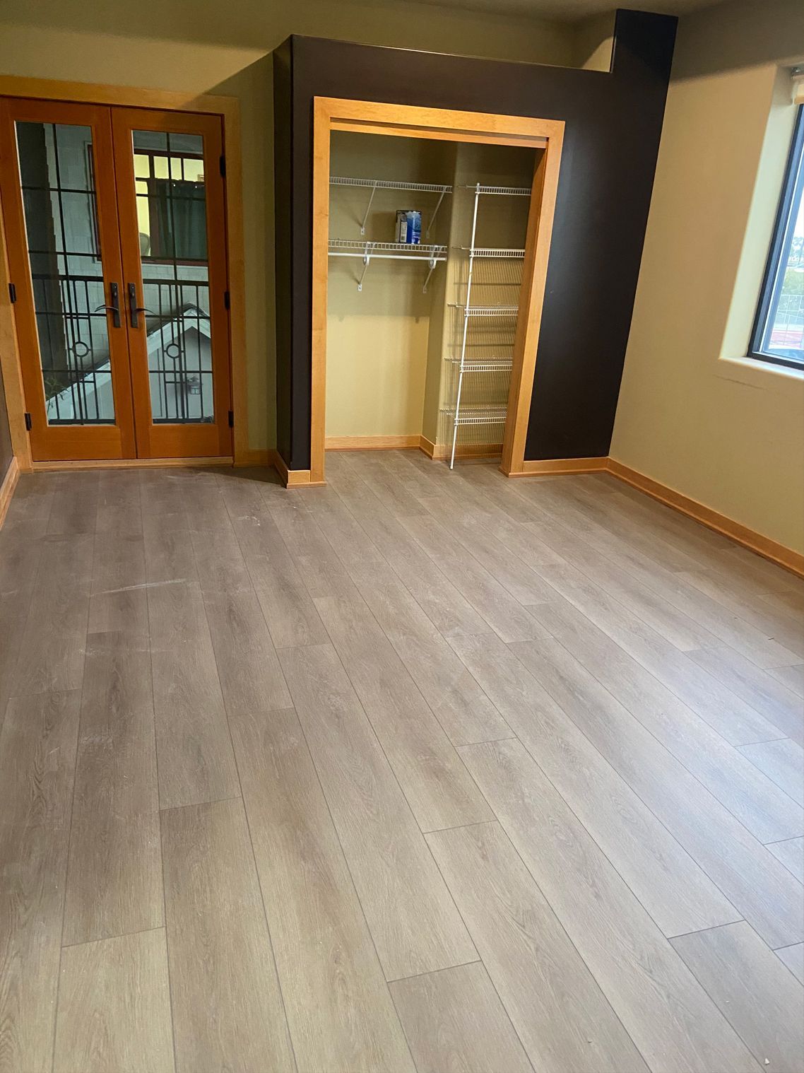 Empty room with gray wood floor, open closet, and wooden double doors near a window.