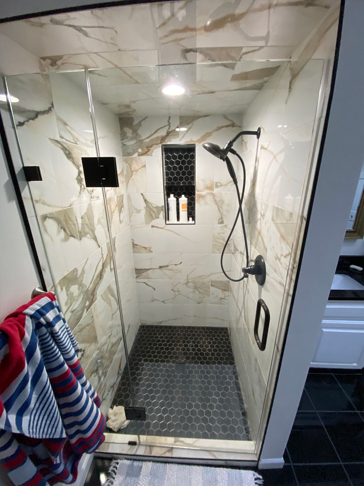 Modern shower stall with marble-look tiles, glass door, black floor mat, and hanging striped towel.