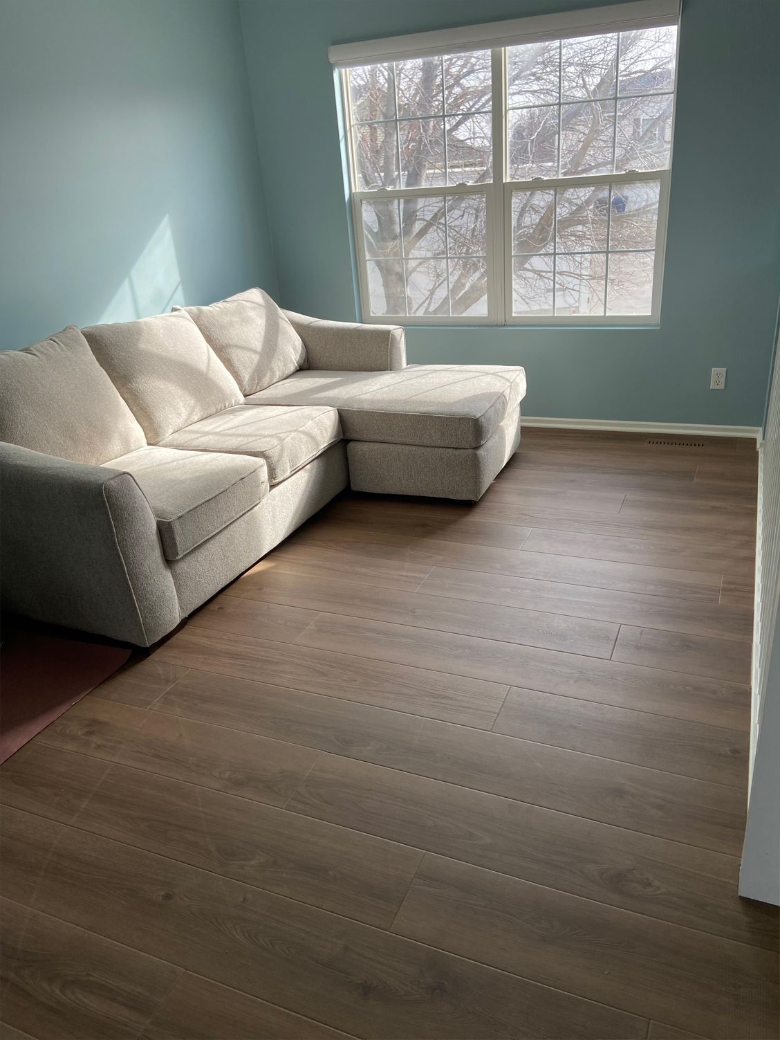 Empty room with a beige sectional sofa, wood floors, pale blue walls, and a large window.