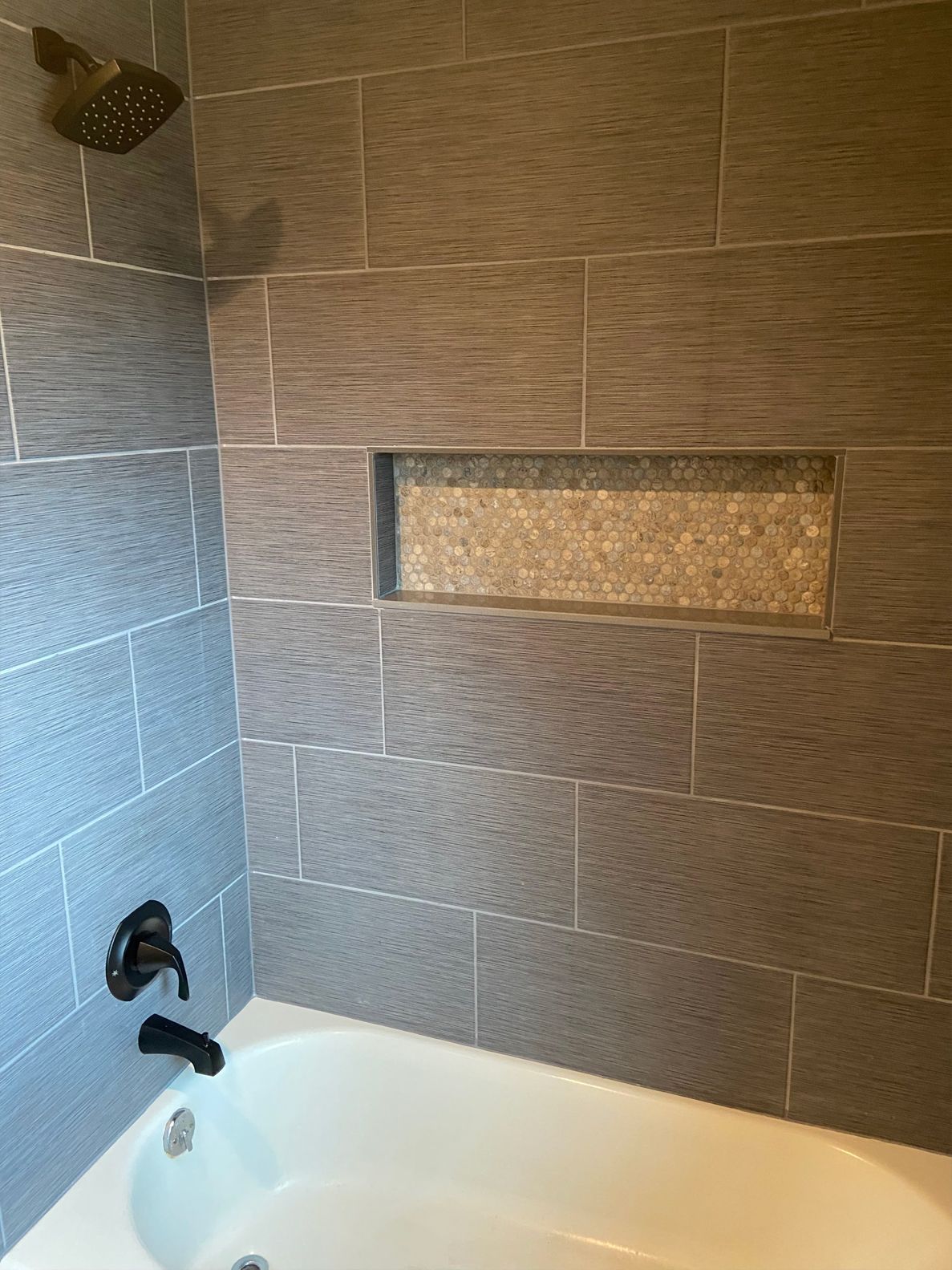 Bathroom shower with gray tile, built-in niche, black fixtures, and part of a white tub visible.