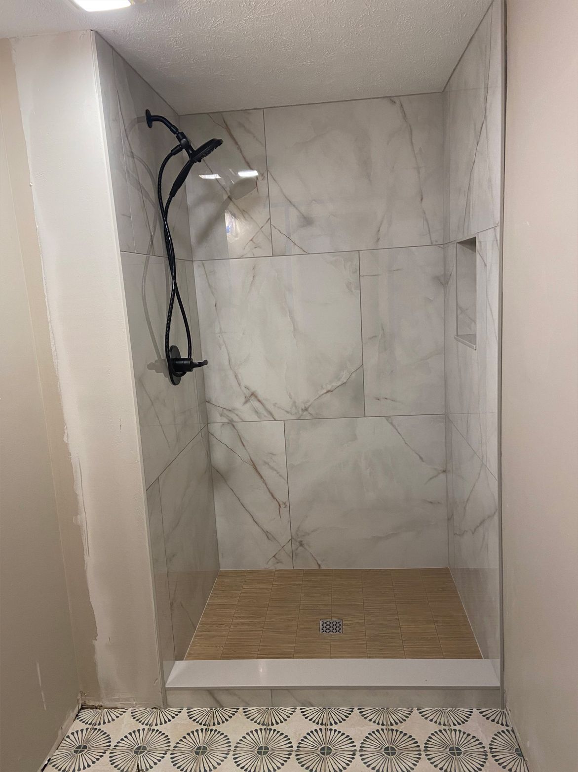 Small tiled shower stall with marble-look walls, black showerhead, and wood-look floor tile.