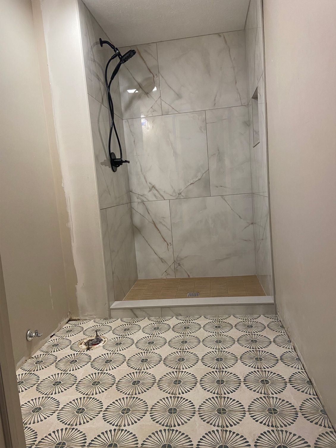 Narrow unfinished shower alcove with marble-look wall tile and patterned tile floor.
