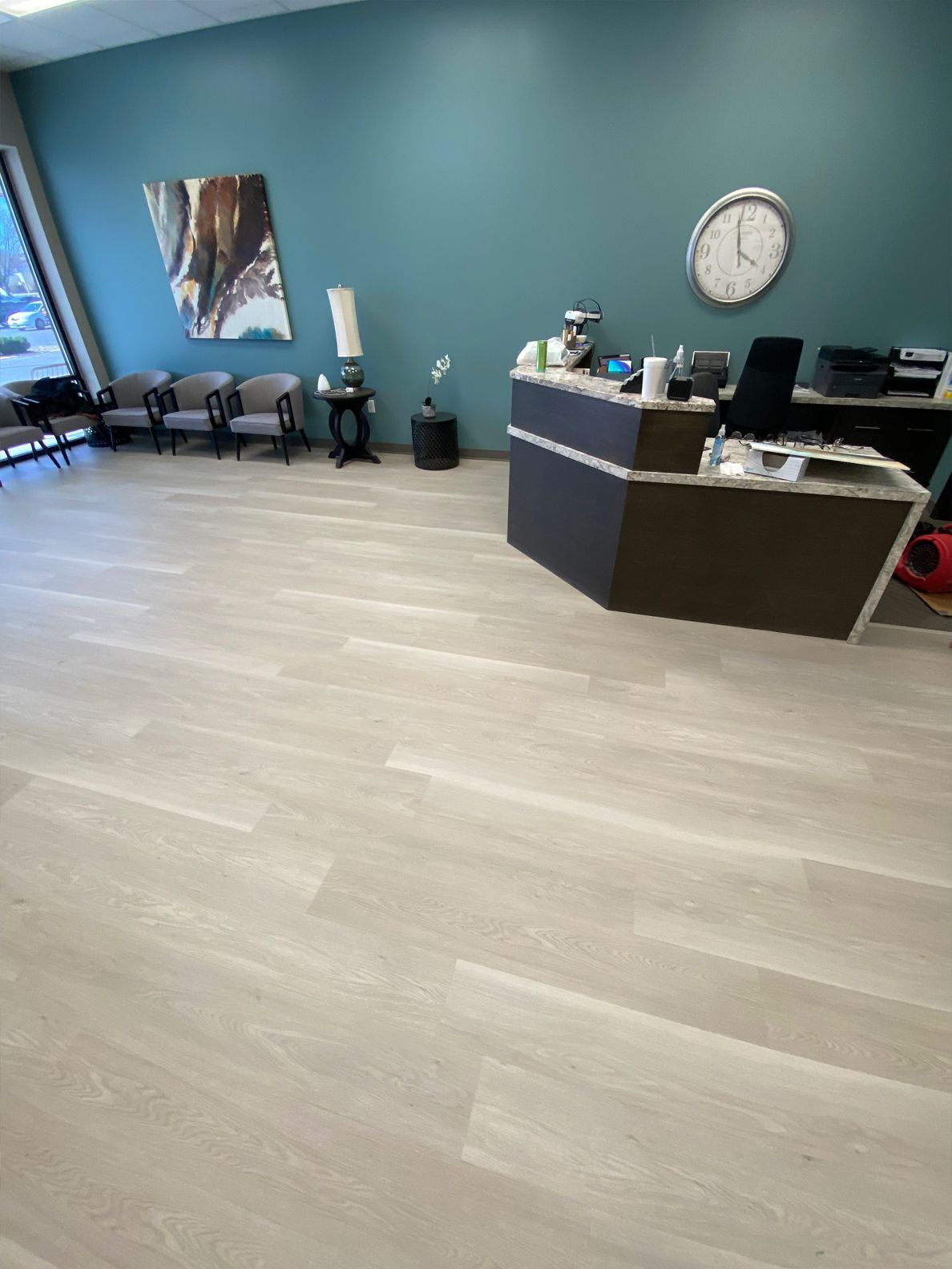 Empty office reception area with teal walls, gray flooring, chairs, and a front desk with a wall clock.