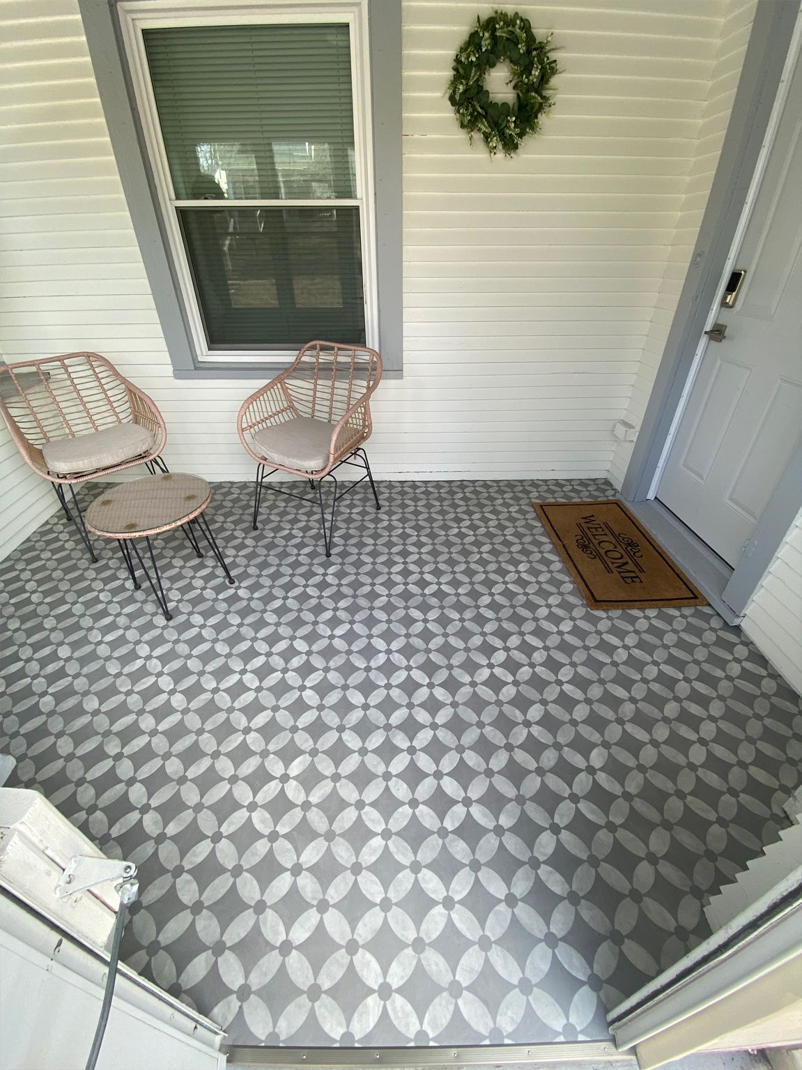 Front porch with two wicker chairs, patterned floor tiles, a wreath, and a doormat beside a white door.