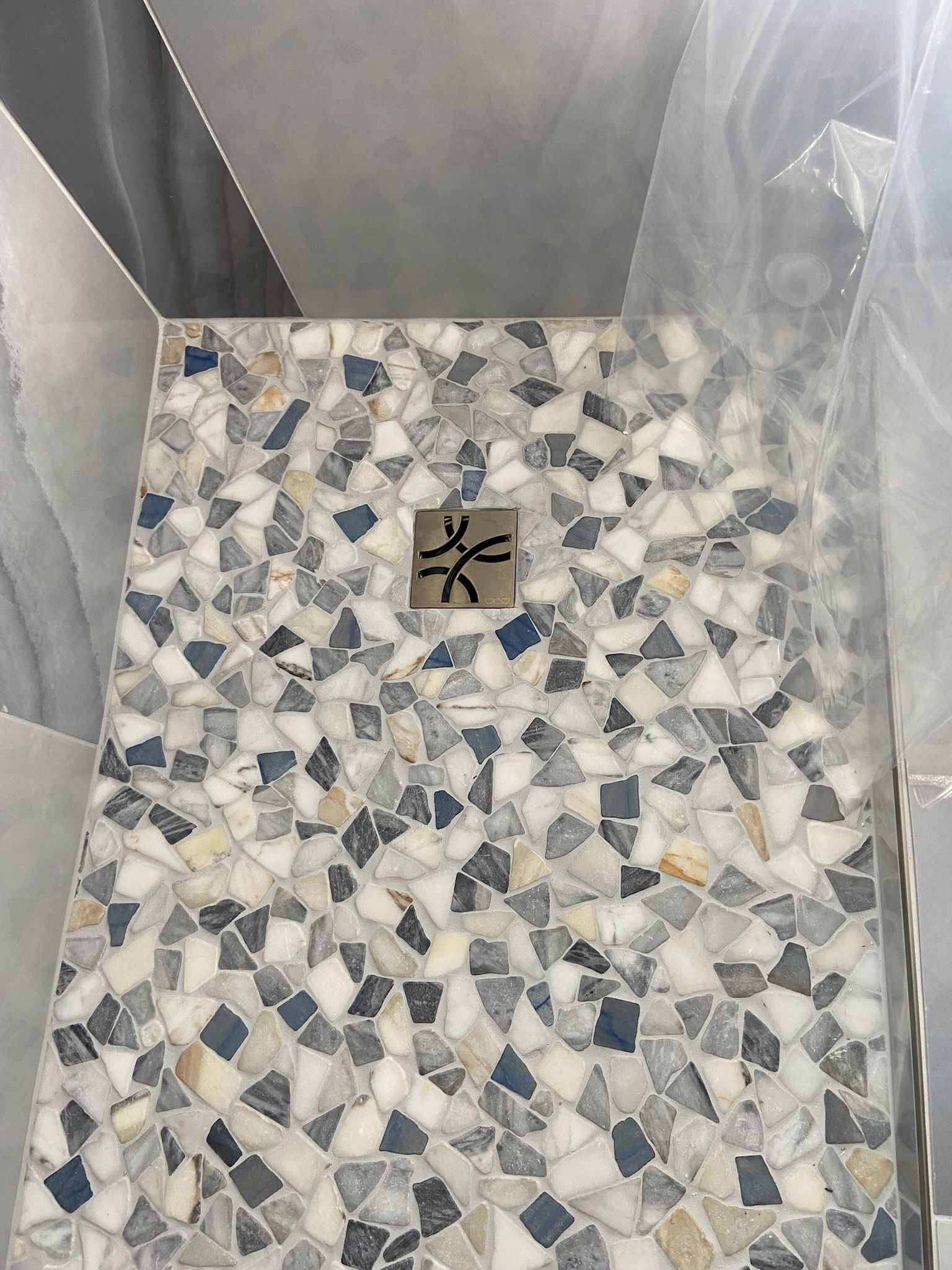 Shower floor with a mosaic tile design in white, gray, and dark blue. A small metal square sits in the center.