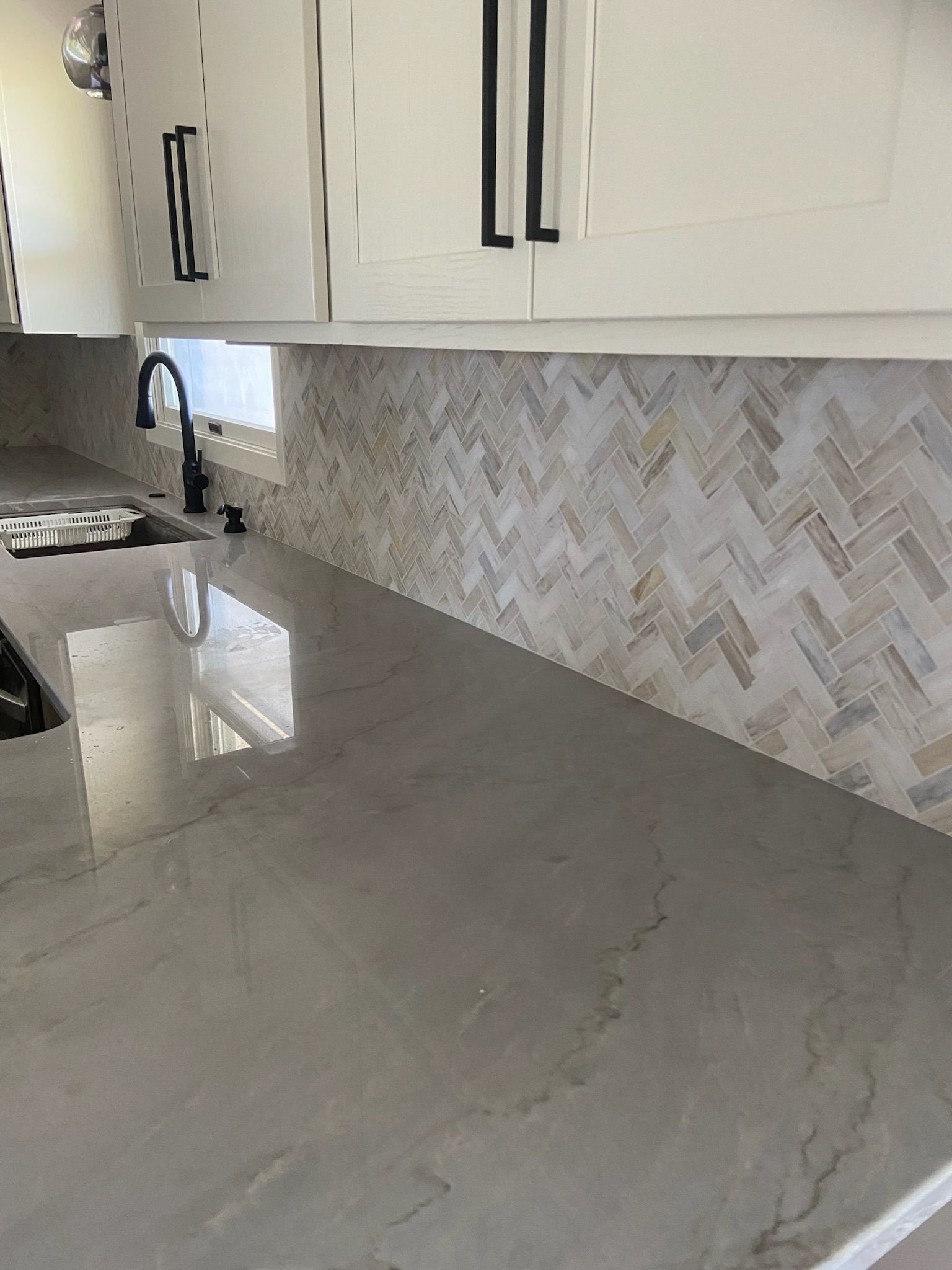 Kitchen countertop with herringbone tile backsplash and white cabinets with black handles.