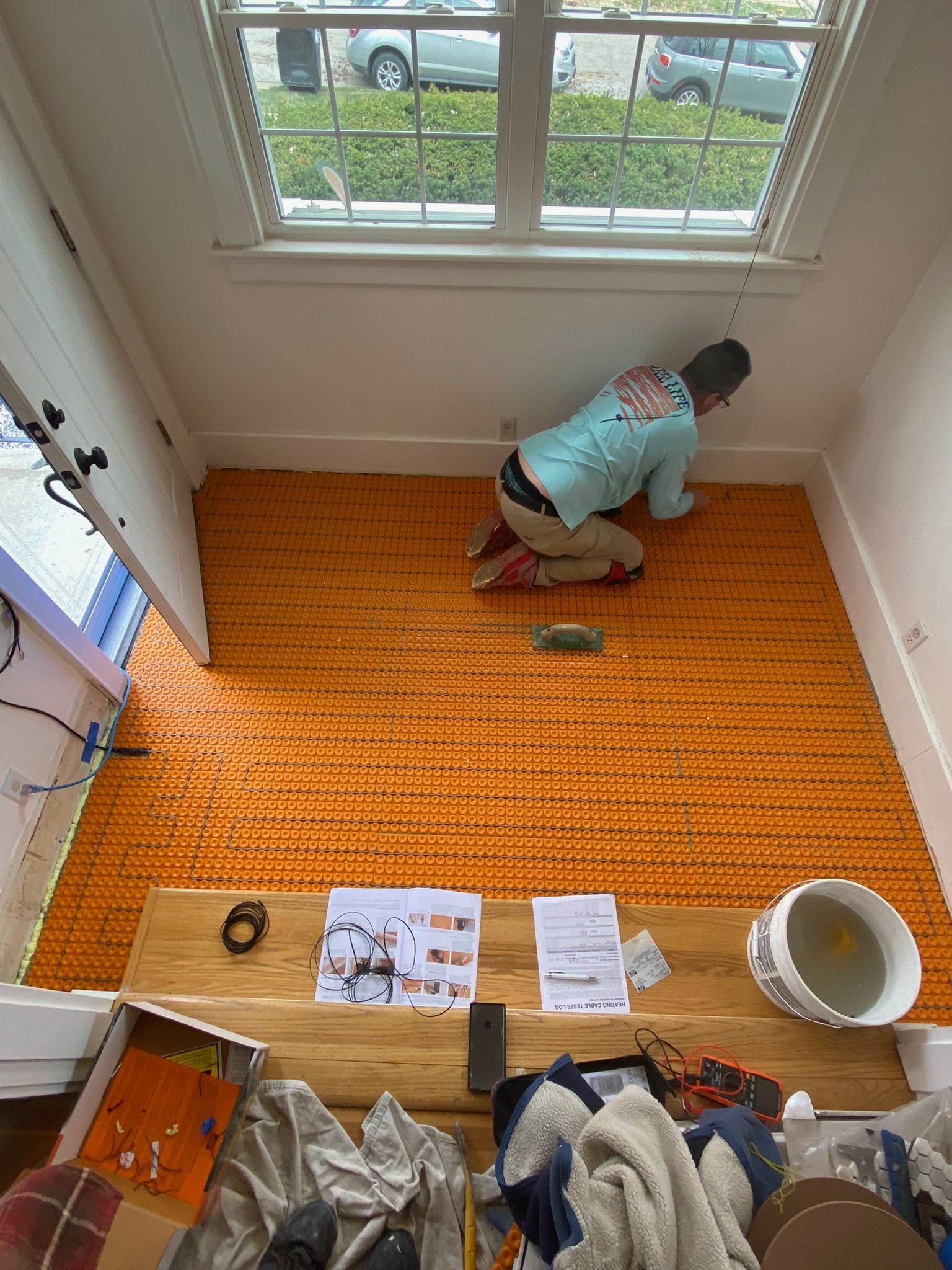 Person installs orange flooring near a window. Tools and debris surround them.