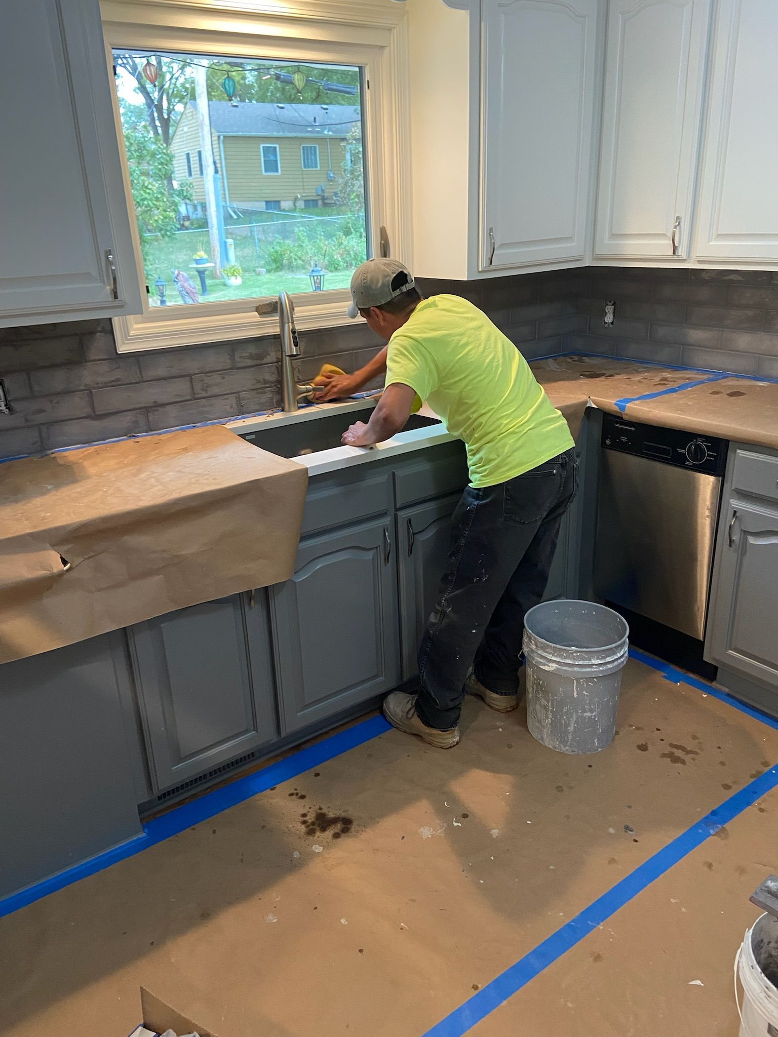 Person sanding a gray kitchen countertop by a sink. Cabinets are gray; walls have tile backsplash.