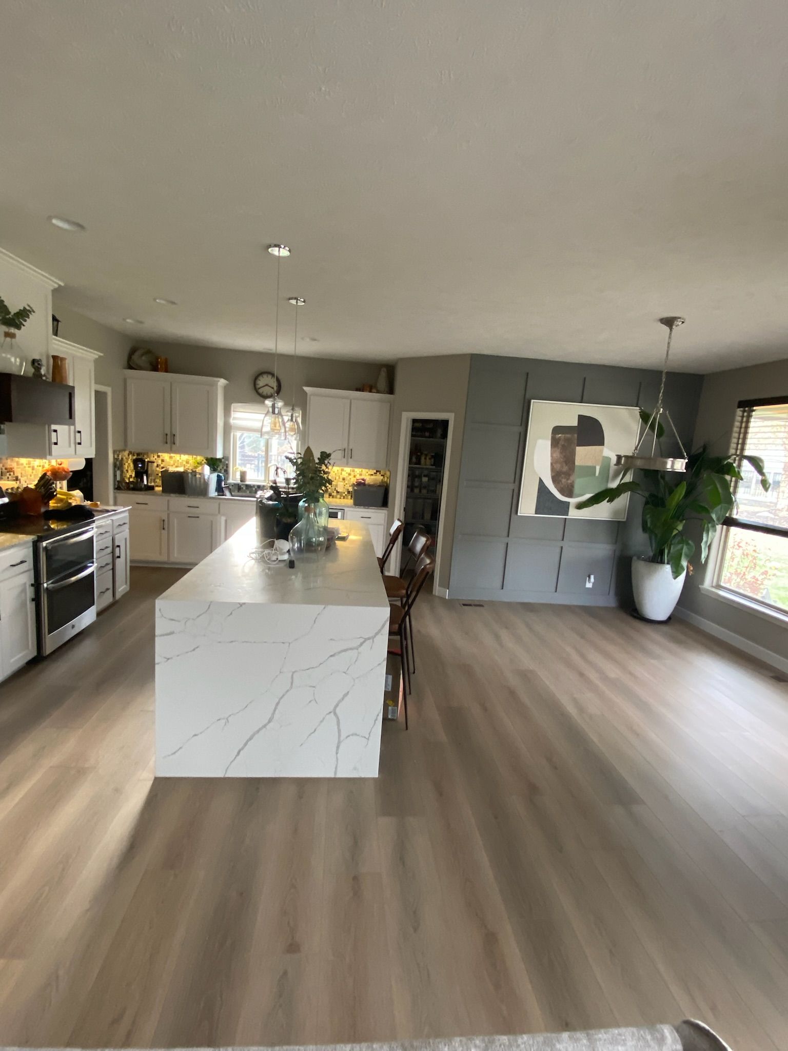 Modern kitchen with large white island, cabinetry, and light wood floors. A large piece of art hangs on a gray accent wall.