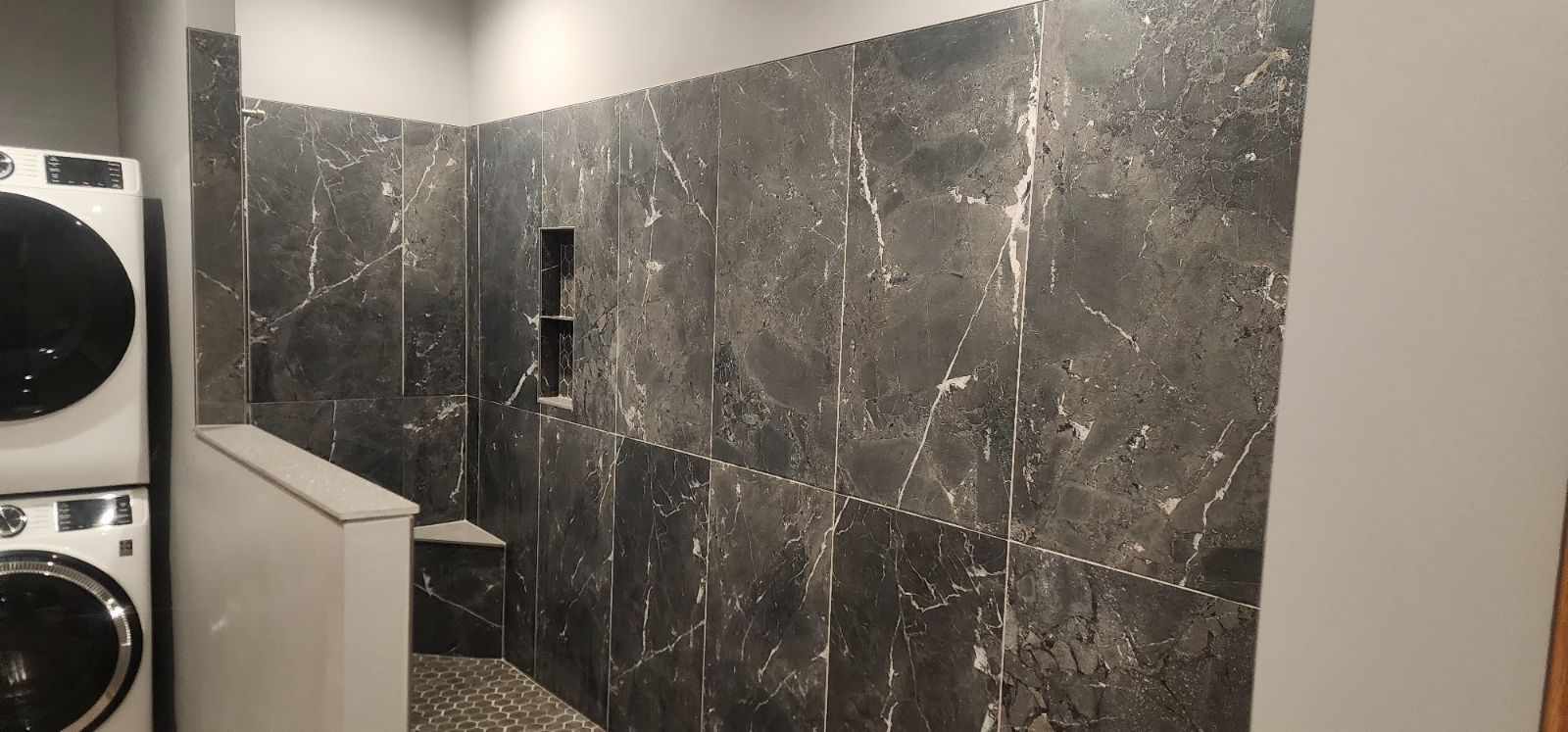 A dark shower stall with black marble-like tiles and a built-in shelf. Next to the shower is a stacked washer and dryer.