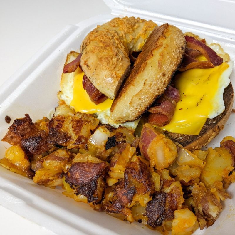 A bagel sandwich with eggs , cheese , bacon and potatoes in a styrofoam container.