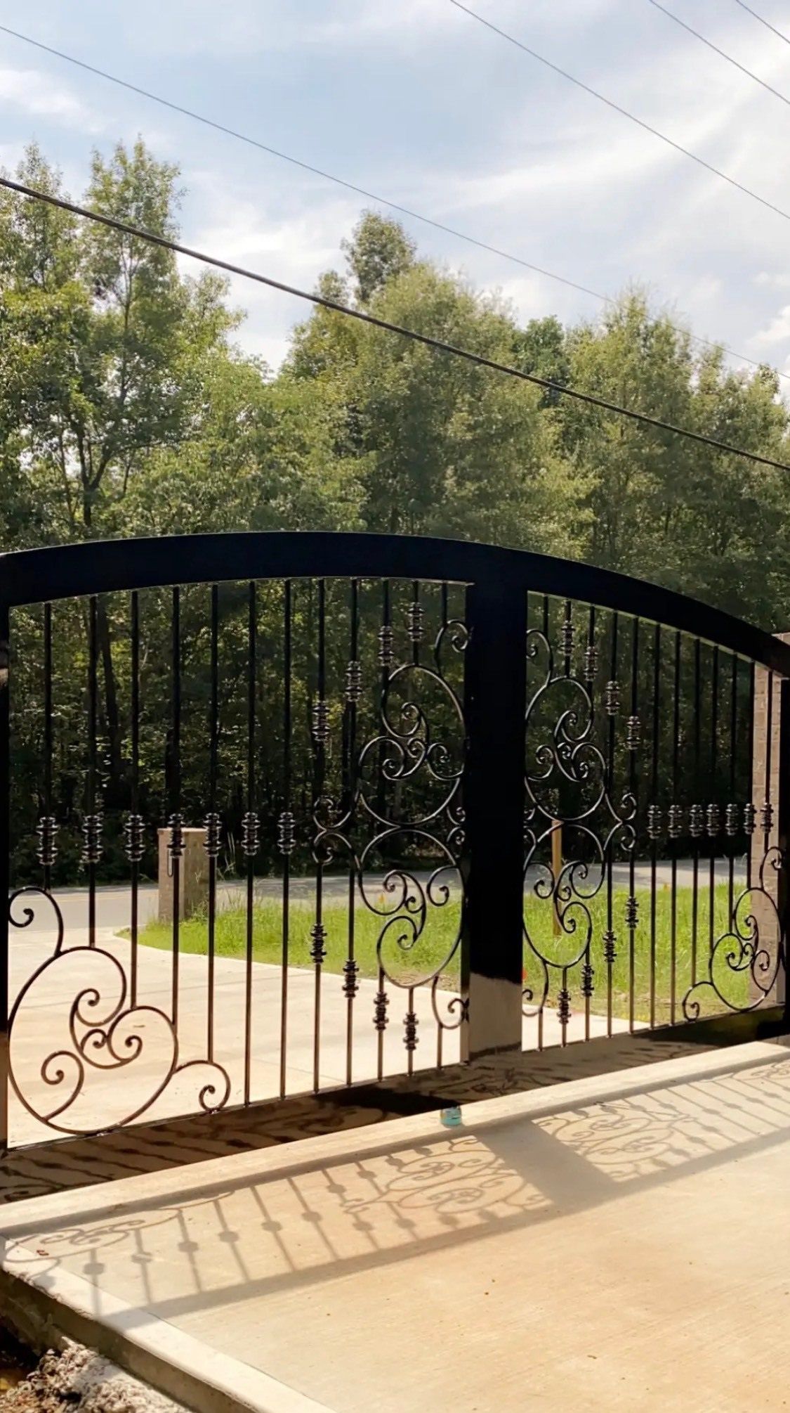 A black wrought iron gate is open to a driveway with trees in the background.