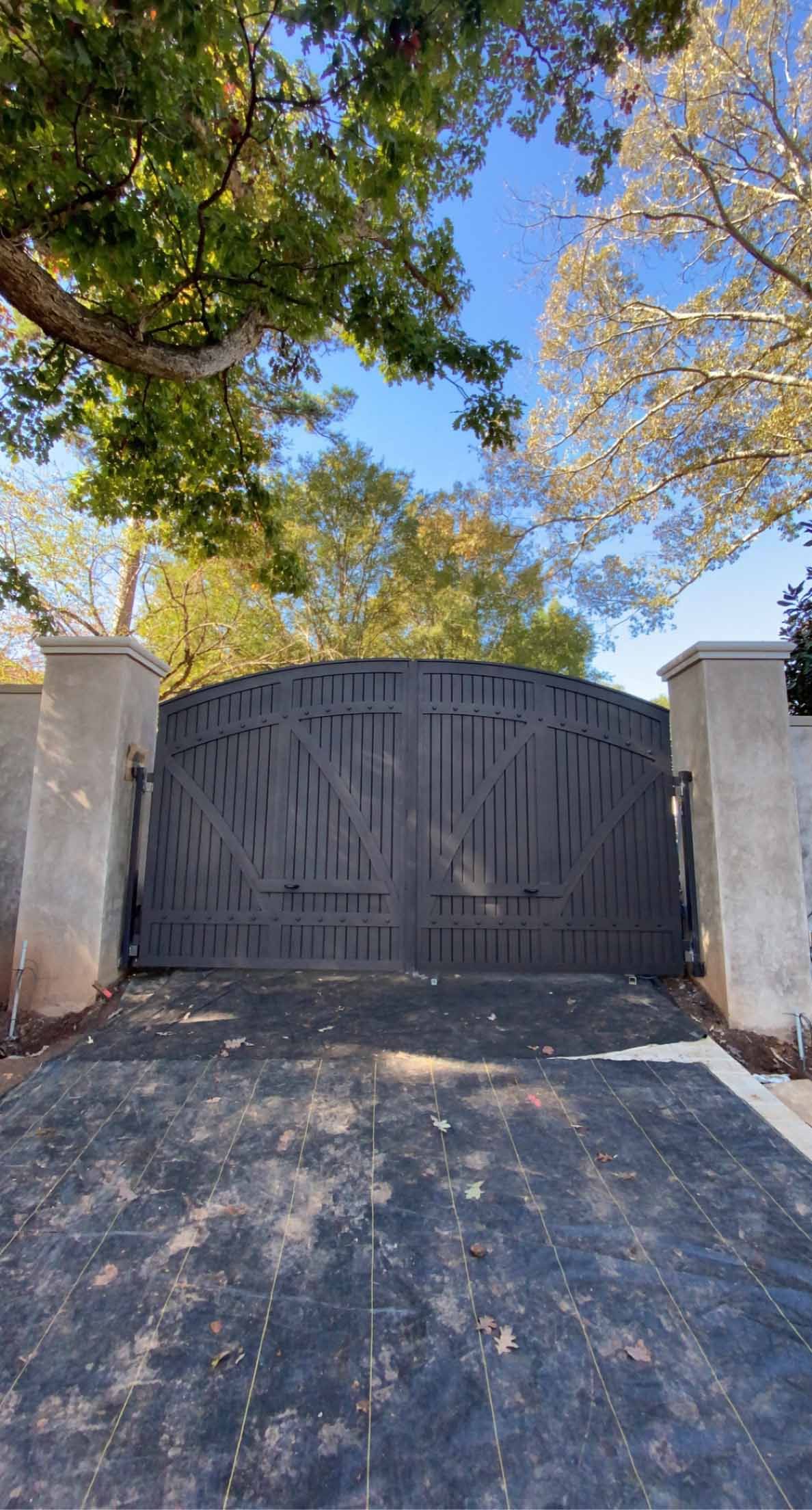 A large metal gate is surrounded by trees and a driveway.