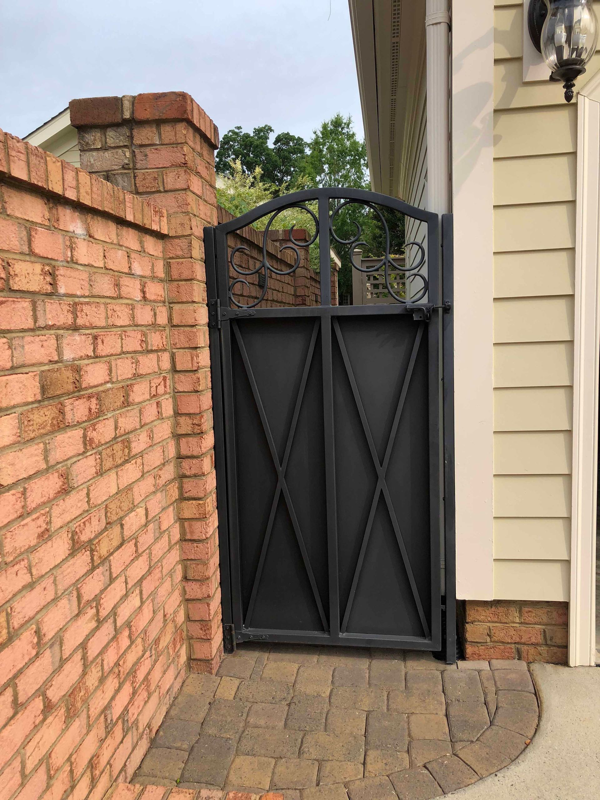 A black gate is between a brick wall and a house.