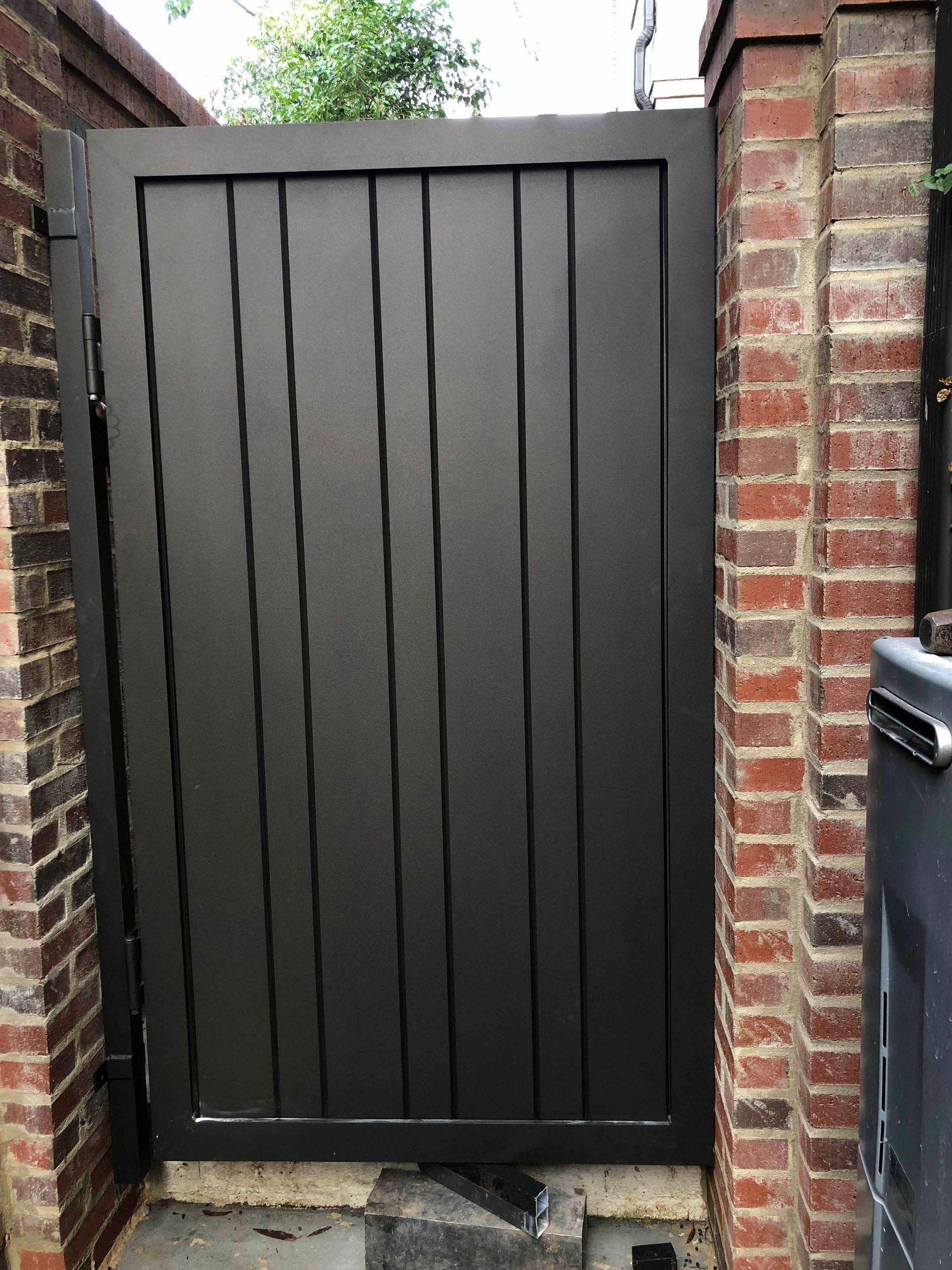A black gate is sitting next to a brick wall.