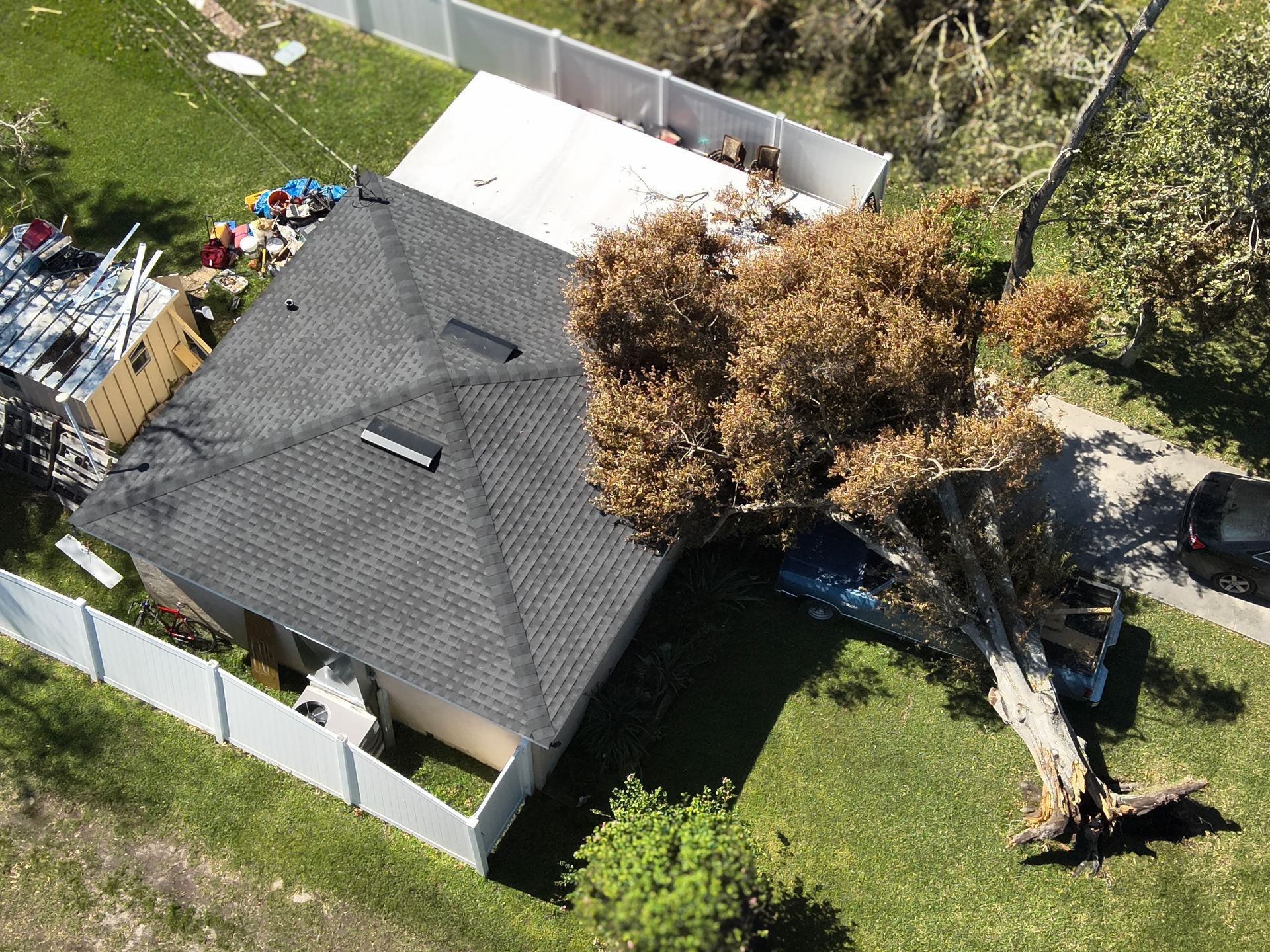 Aerial view of a house damaged by a fallen tree; car crushed, debris visible, white fence, green lawn.