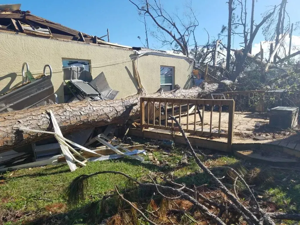 A house with damaged roof and a tree fallen across the deck and lawn after a storm.