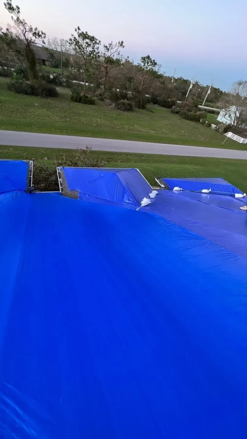 Blue tarp covers a structure on a grassy area, trees and road in the background.