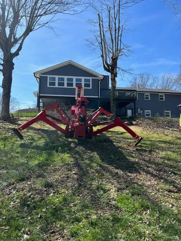 Red spider-like tree trimming machine on a grassy hill in front of a blue house under a blue sky.
