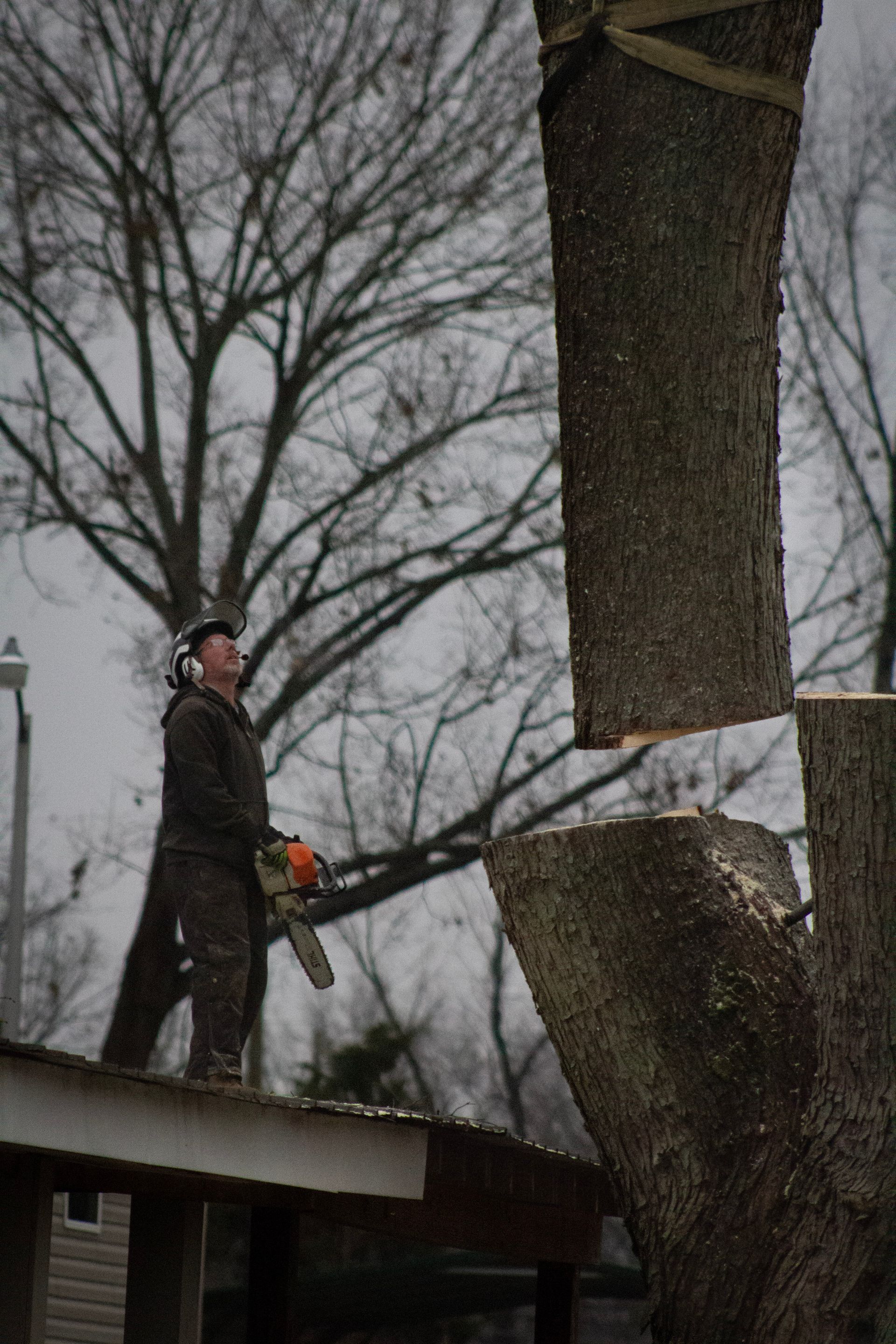 Man using a chainsaw to cut a tree trunk section. He stands on a rooftop, with a cloudy sky in the background.