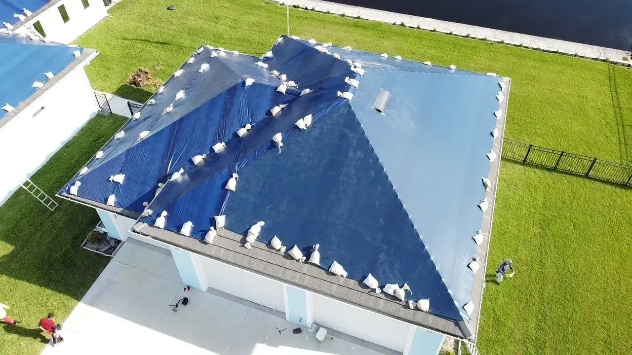 Aerial view of a blue-tarped roof with white anchors, covering a house with a green lawn.