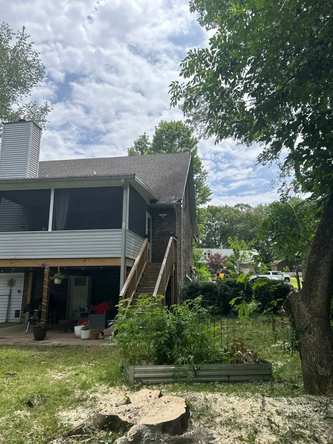 Back view of a light blue house with a screened porch and stairs, next to a garden and tree under a cloudy sky.