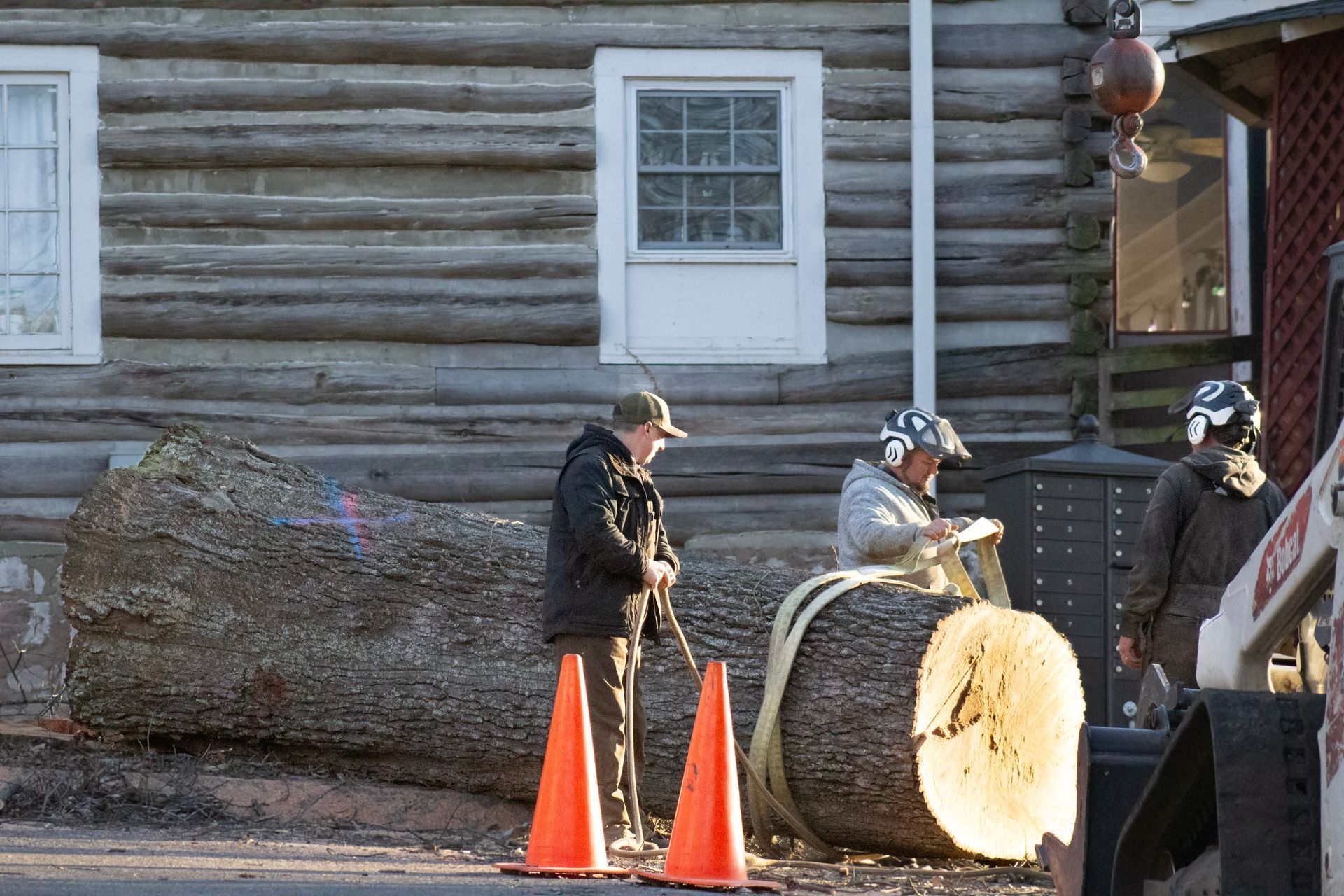 Men using chainsaws to cut a large log near a log cabin; safety cones present.