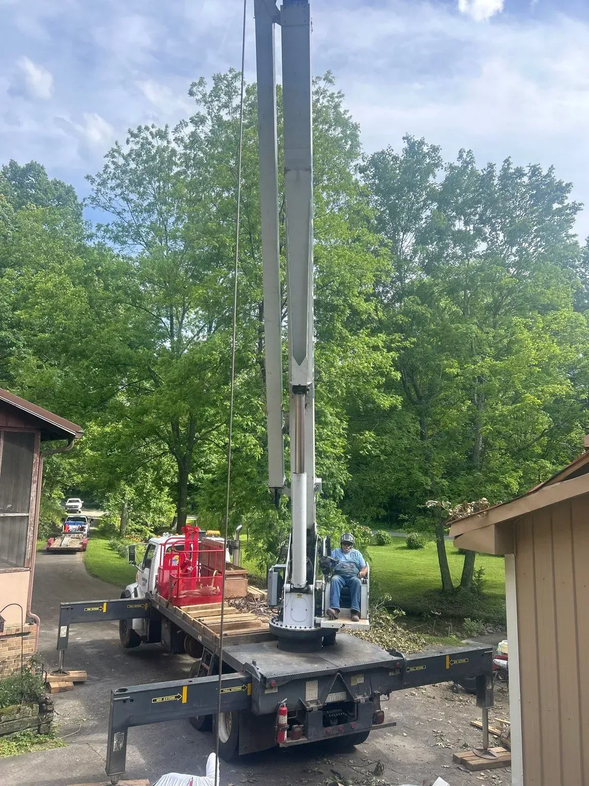 A truck with a crane extending upwards. Two people are working, with lush green trees in the background.
