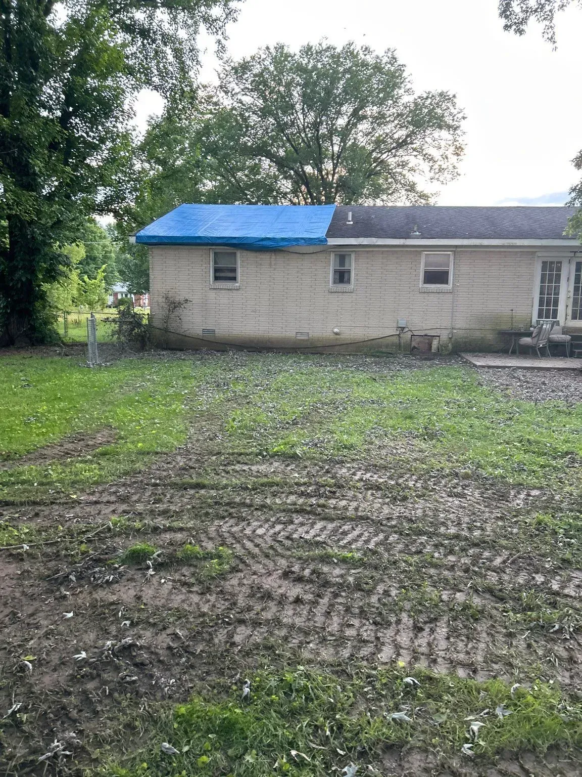 Backyard with house; blue tarp on roof. Muddy yard with tire tracks, grass, and trees.