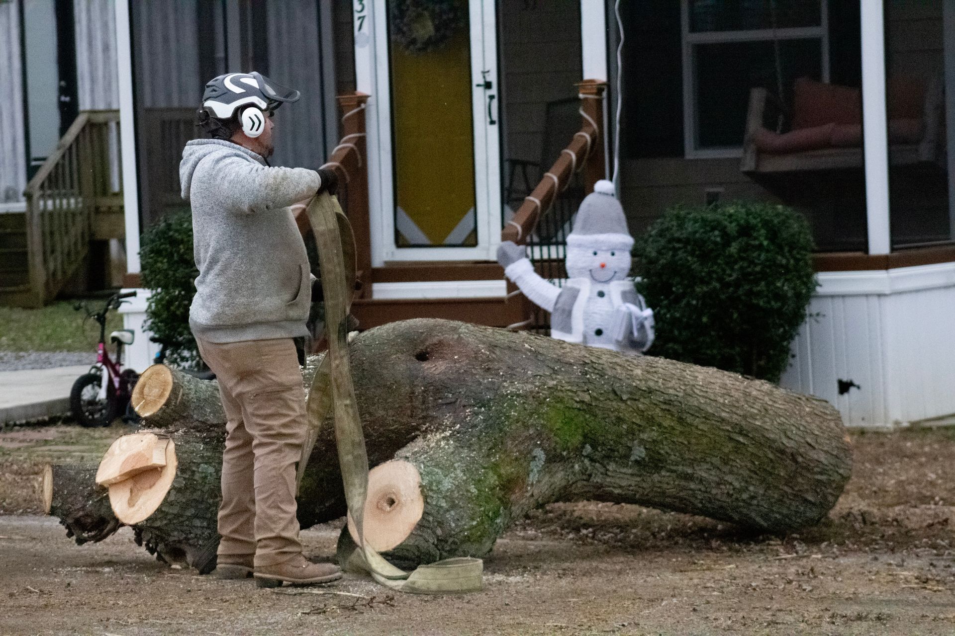 Man sawing a large log with safety gear in front of a house decorated with a snowman and holiday lights.
