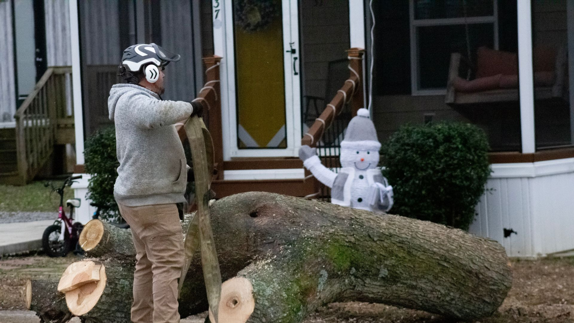 Person sawing a large log with safety gear in front of a house. A snowman decoration is nearby.