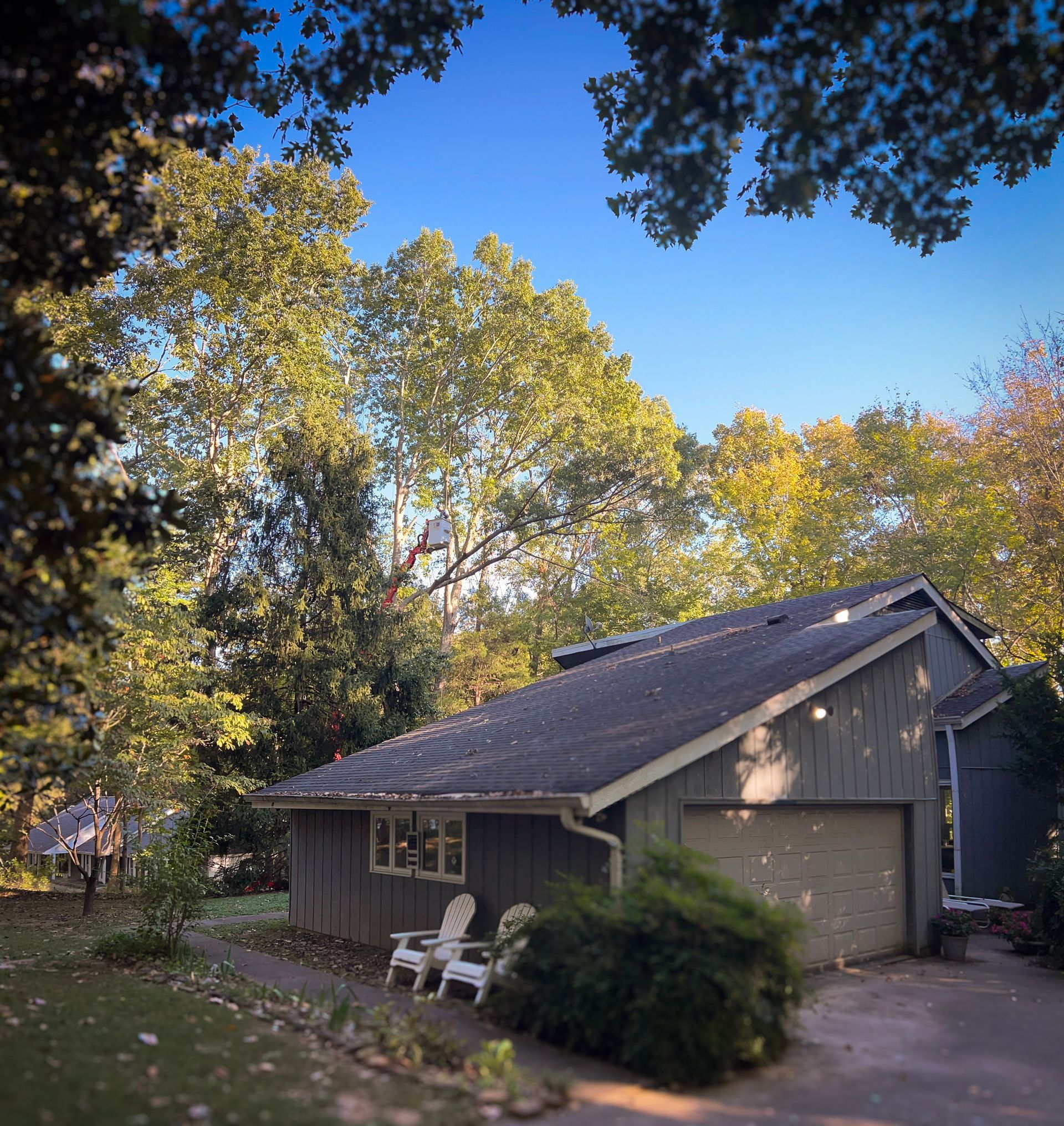 A gray house with a garage and two chairs in a yard under a blue sky and trees.