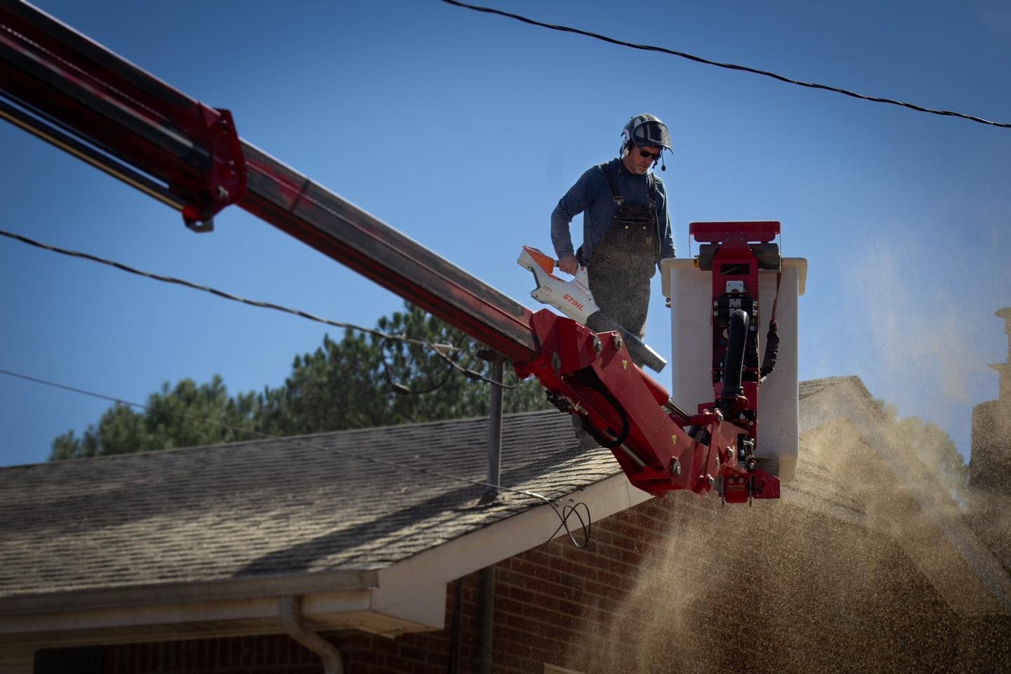 Worker in a red bucket truck trimming a tree, debris falling. Bright blue sky, residential setting.