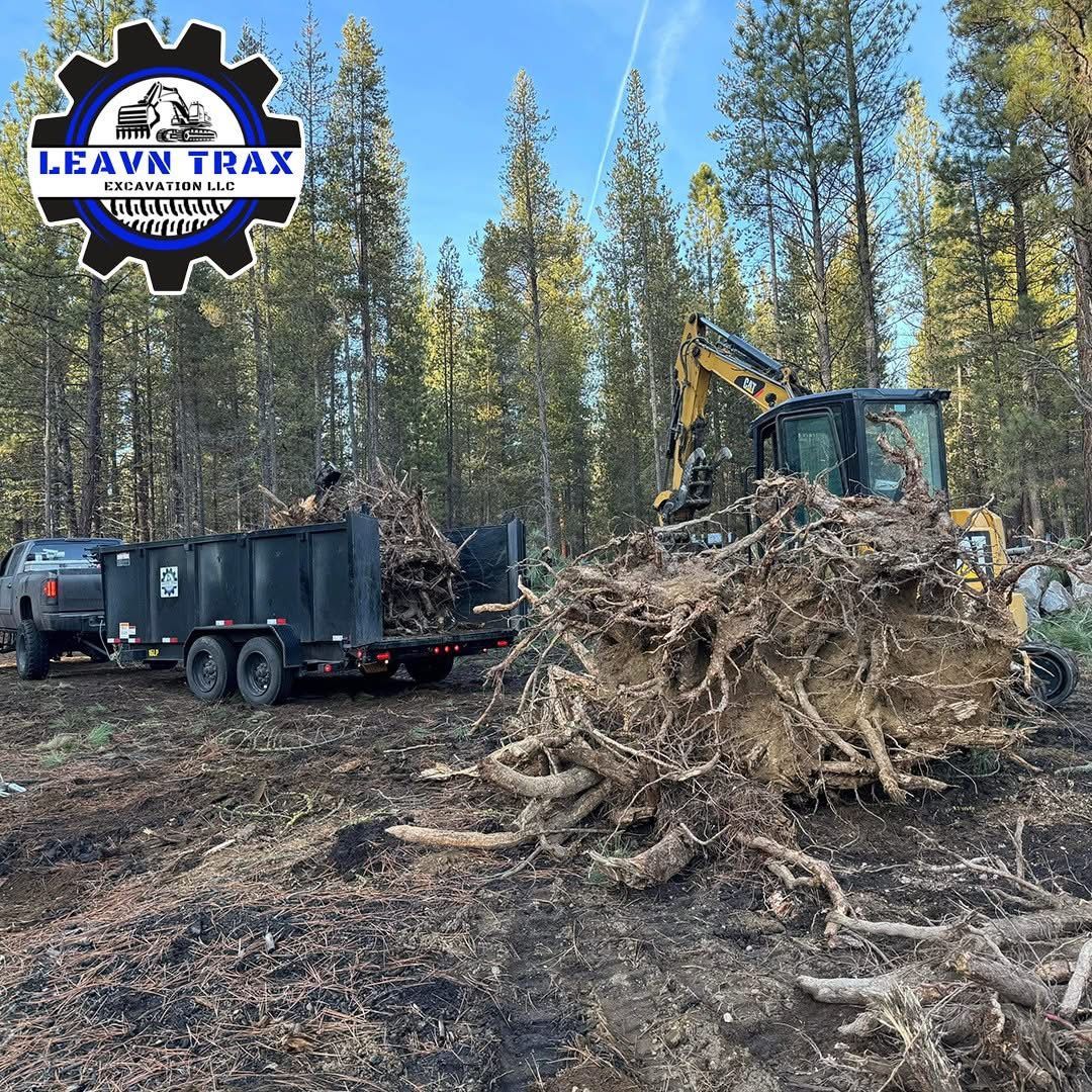 Excavator and trailer clearing uprooted tree roots in a forest, with a LEAVN TRAX logo in the corner