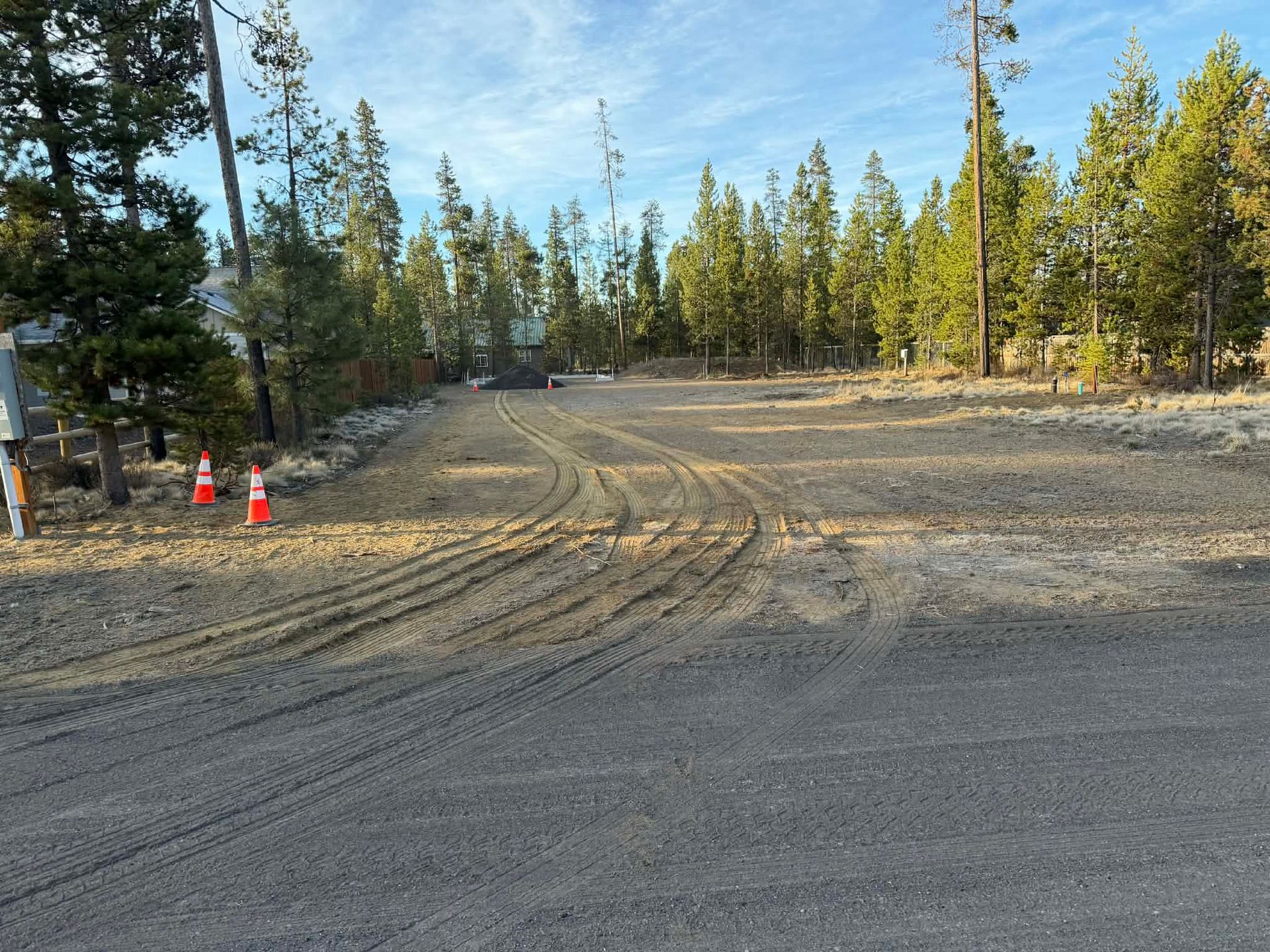 Gravel road under construction beside a forest, with orange cones and tire tracks in the dirt.