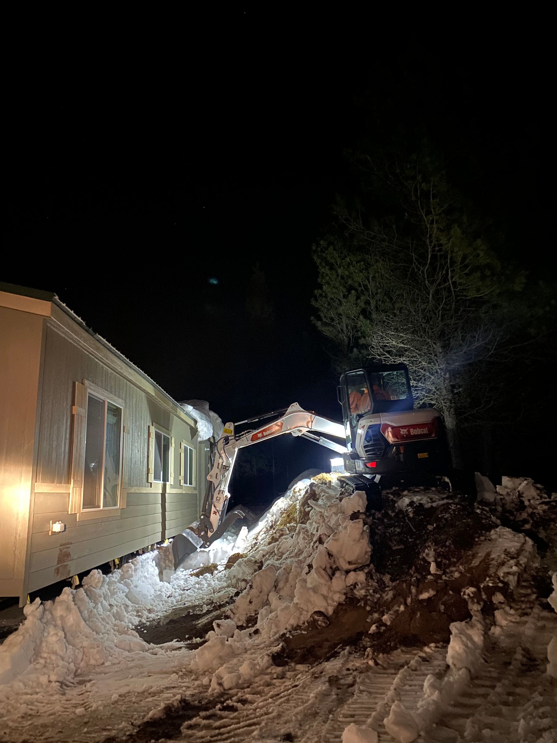 An excavator is clearing snow from a house at night.