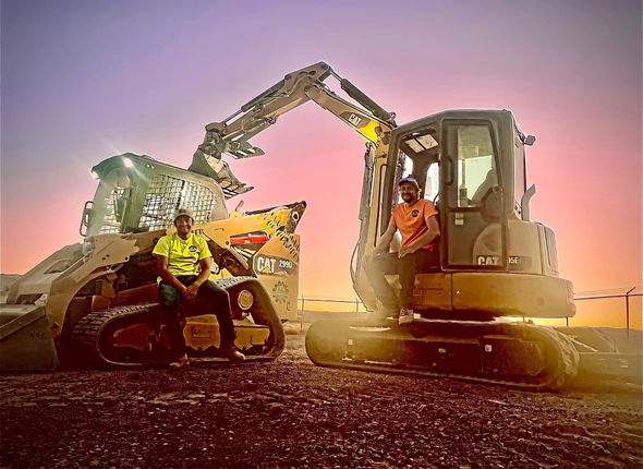 Two men are sitting on a bulldozer with a sunset in the background.