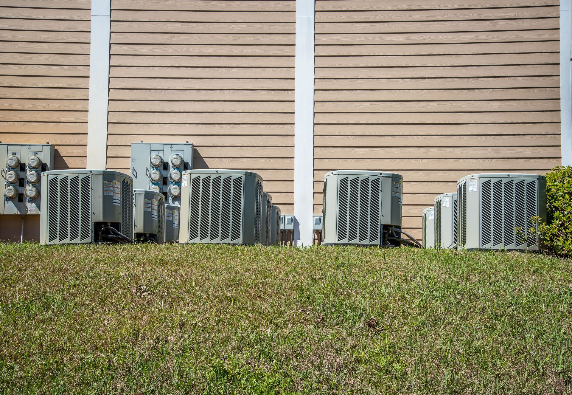 Outdoor HVAC units lined up against a tan building wall behind a grassy lawn.