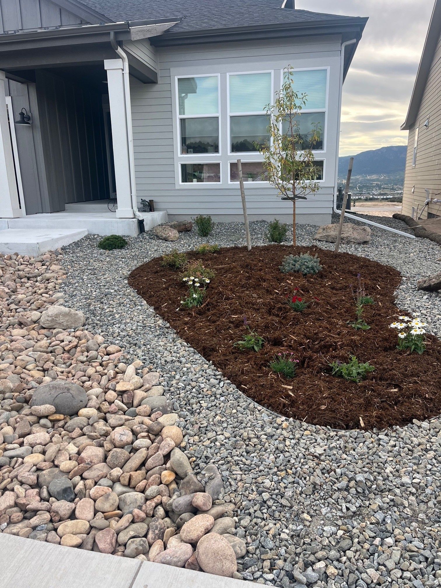 Front yard landscaping with gray rocks, mulch, and small plants in front of a light gray house.