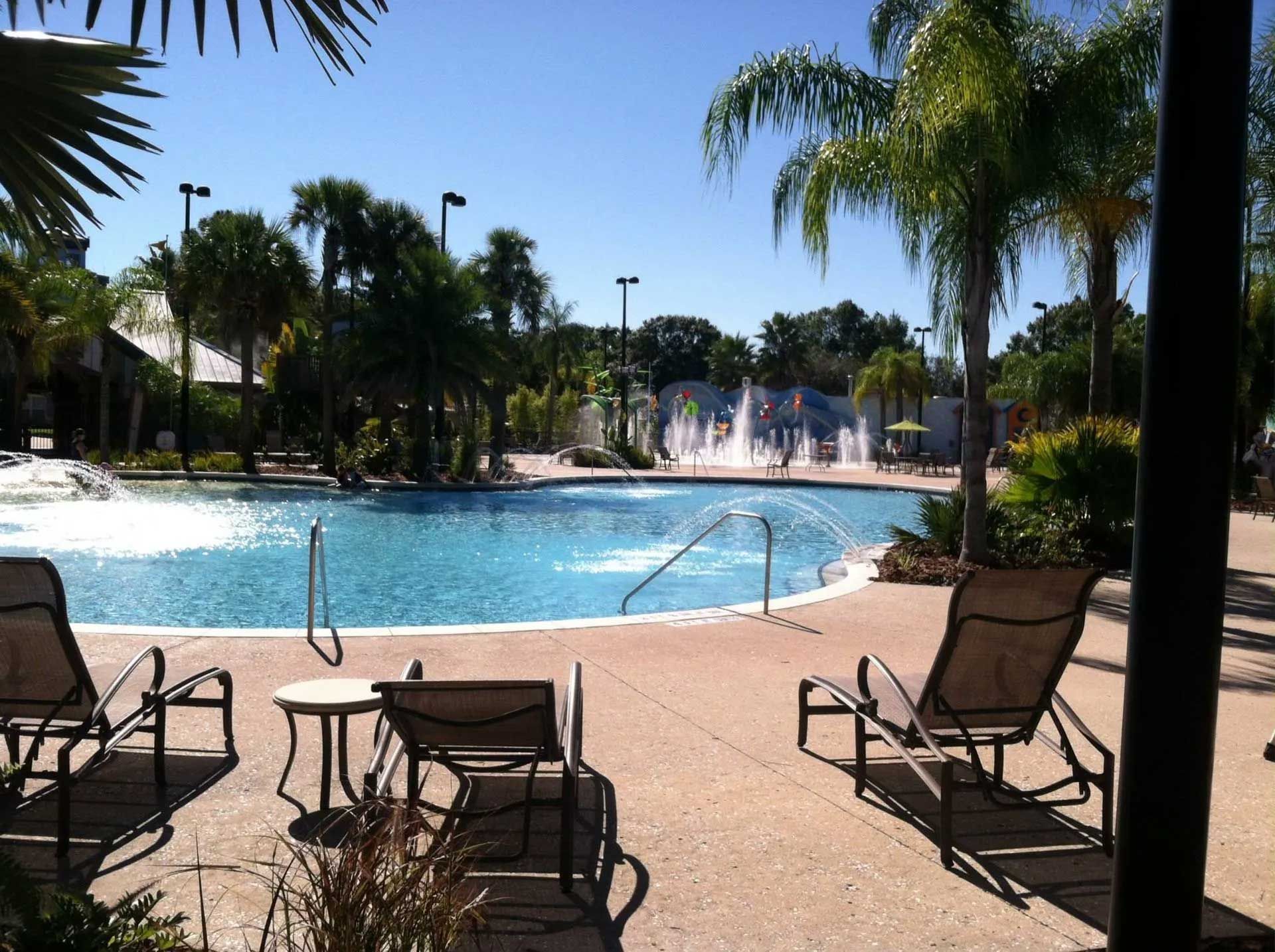 A sunny outdoor pool with lounge chairs, palm trees, and fountains in the distance.