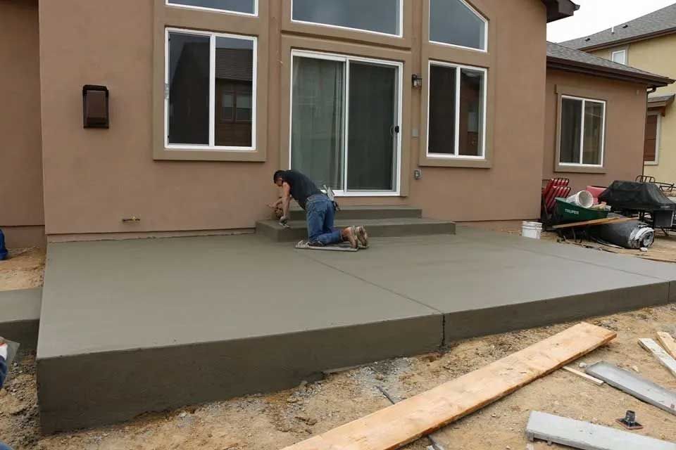 Man smoothing wet concrete patio near a house with sliding glass doors; overcast day.