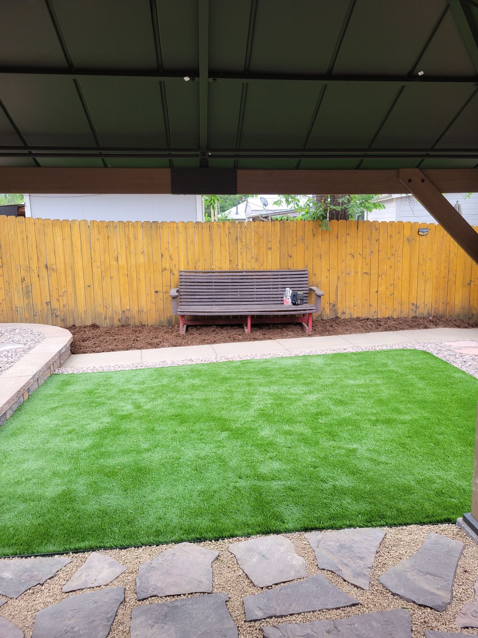 A backyard with artificial turf, a bench, and a wooden fence, viewed from under a patio roof.