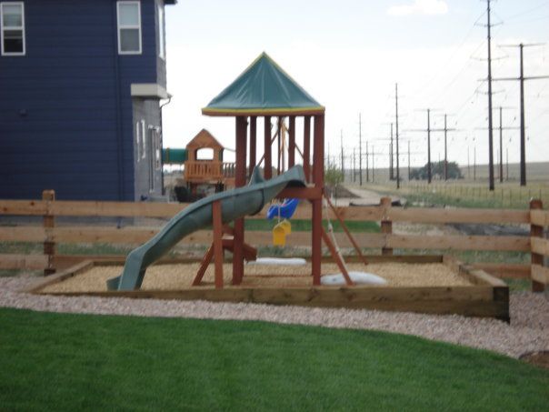 Playground with a slide, sandbox, and swingset in a yard with a wooden fence.