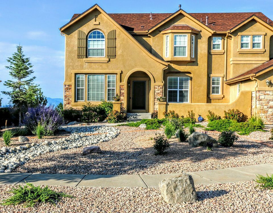 Tan stucco house with brown roof, shutters, and a gravel front yard.