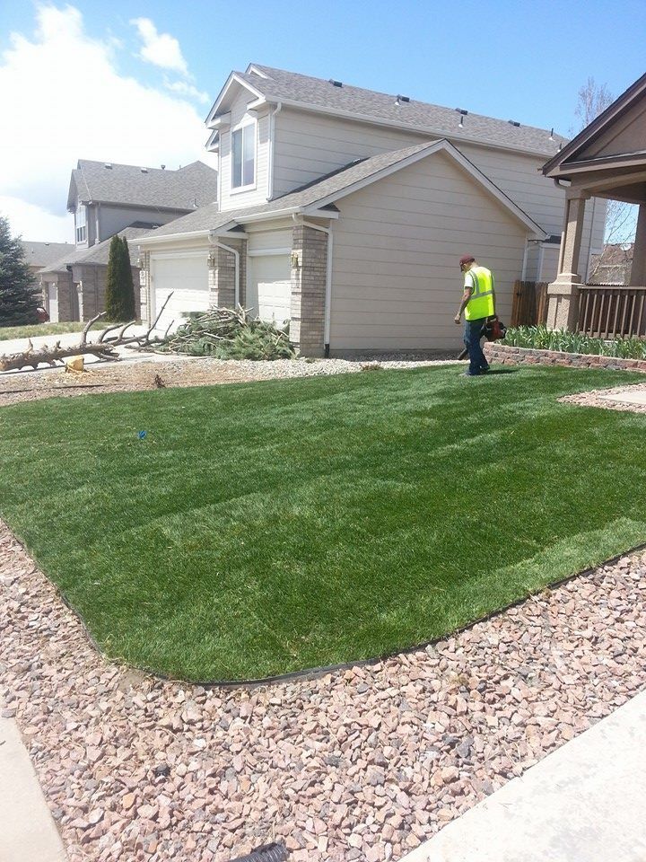 A person in a yellow vest mows green grass in front of a two-story beige house.