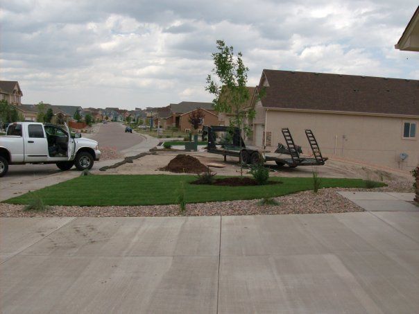 A white truck parked on a driveway with landscaping, a trailer, and a house on a suburban street.