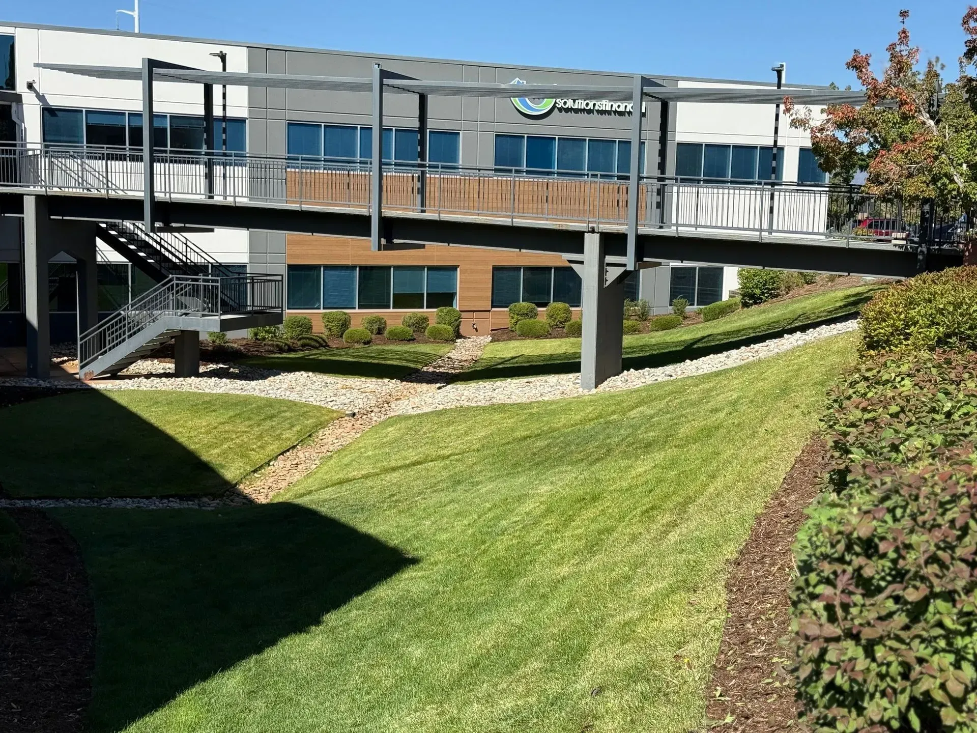 A modern office building with a walkway bridge, grassy hills, and blue sky.