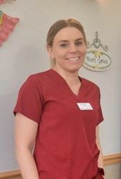 A woman in a red scrub top is smiling and standing in front of a white board.