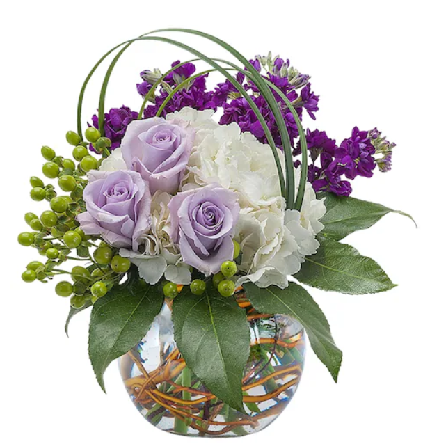 Floral arrangement in a glass bowl; lavender roses, white hydrangea, and purple stock with green accents and foliage.